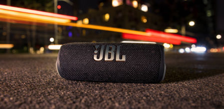 Altavoz Bluetooth JBL portátil sobre el asfalto por la noche con luces de ciudad desenfocadas al fondo