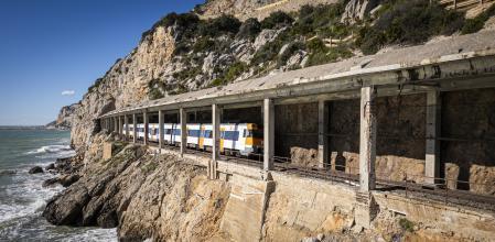 Un tren de Rodalies circula por la línea del Garraf bajo una estructura visiblemente maltrecha