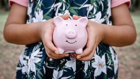 Close-Up Portrait of Little Girl Holding Piggy Bank Against Backyard Garden Background