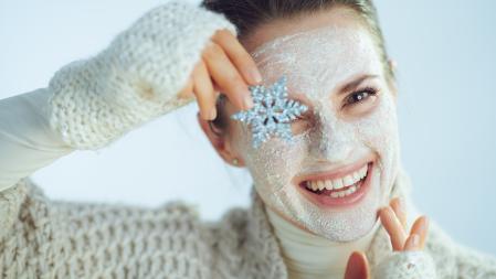 Portrait of smiling modern middle age woman in roll neck sweater and cardigan with white facial mask showing snowflake on winter light blue background. (Portrait of smiling modern middle age woman in roll neck sweater and cardigan with white facial ma