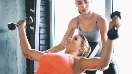 Closeup of two women at a gym. One of them is working out with dumbbells by stretching and flexing her pecs and the other one is her spotter. Front view.
