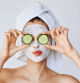 Mujer joven con una mascarilla facial sujetando dos trozos de pepino en sus ojos
