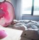 A young girl sleeps in her bed and has a pink fan on her desk blowing cool air over her.