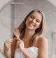 Haircare concept. Pretty lady brushing her beautiful hair with comb while looking at her reflection and smiling in mirror in bathroom interior