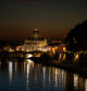 La majestuosa cúpula de la Basílica de San Pedro, iluminada y dominando el horizonte, mientras que el puente que cruza el Tíber se refleja en las aguas tranquilas del río.