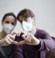 couple with face mask doing a heart with their hands while sitting on the couch