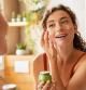 Woman caring of her beautiful skin face standing near mirror in the bathroom. Young woman applying moisturizing cream on her face during morning routine. Smiling natural girl holding little green jar of ecological cosmetic cream.
