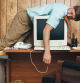 A man and office in 1980's - 1990's style, complete with vintage computer and technology of the time, sleeps on top of his desk, slumping over the computer, too tired and bored to continue working.  Wood paneling on the wall in the background.