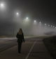 Young woman alone slowly walking under white street lights in night. Dark time. Peaceful atmosphere in mist. Foggy air. Back view.