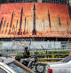 A motorcycle speaks on a phone while driving past a billboard depicting named Iranian ballistic missiles in service, with text in Arabic reading 