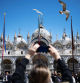 A woman uses her smartphone to take footage of the Basilica with flying gulls in San Marco Square in Venice, on April 25, 2024. The new strategy to lower the number of tourists visiting the UNESCO World Heritage site calls for day-trippers to pay a five-euro ticket to enter the historic city centre and is due to start on April 25. (Photo by MARCO BERTORELLO / AFP)