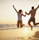 Rear view of a happy young couple jumping on the beach while holding hands at sunset