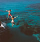 Young active man and woman diving from high cliff into tropical island blue sea water