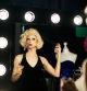 A drag queen prepares for a performance backstage, applying hairspray in front of a mirror with bright lights.
