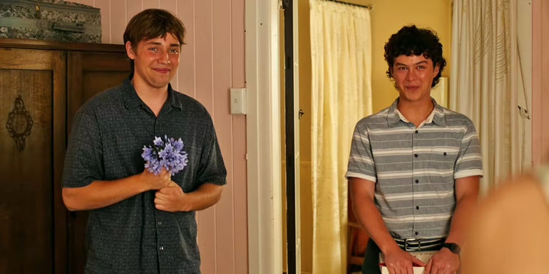 Lee Tiger Hailey as Gus Bell holding light purple flowers and Zac Burgess as Older Eli Bell holding thin white gift box by a doorway in Boy Swallows Universe