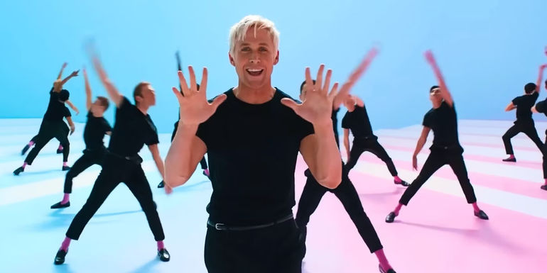 Ryan Gosling's Ken in Barbie standing and smiling while holding up ten fingers. The background is a blue wall and a blue and pink floor, and various other Kens, dressed in black just like Gosling, are dancing in the background.