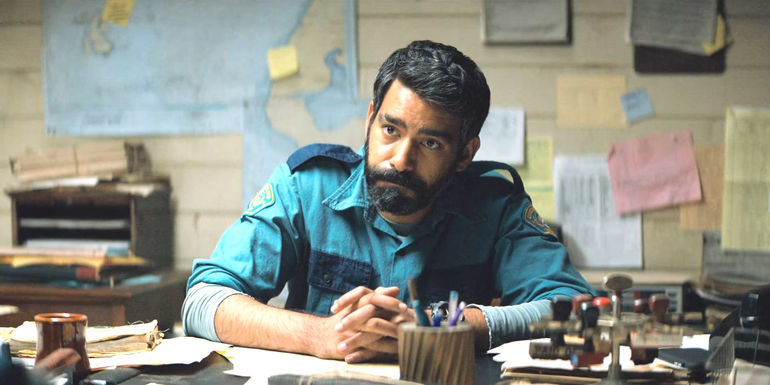 Rahul Kohli in Midnight Mass in a police uniform sitting behind his cluttered desk attentively listening to someone speaking