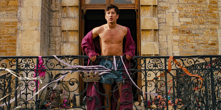 Barry Keoghan as Oliver standing at a balcony covered in streamers wearing a bathrobe and boxers in Saltburn