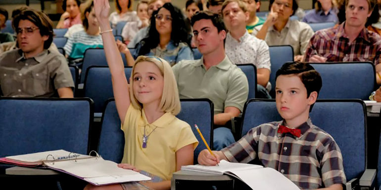 Paige (Mckenna Grace) and Sheldon (Iain Armitage) in class surrounded by other students in Young Sheldon, with Paige enthusiastically raising her hand.
