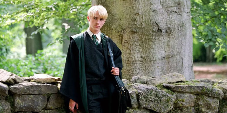 Draco Malfoy against a stone wall with a bag over his shoulder outside of Hogwarts in the Harry Potter movies