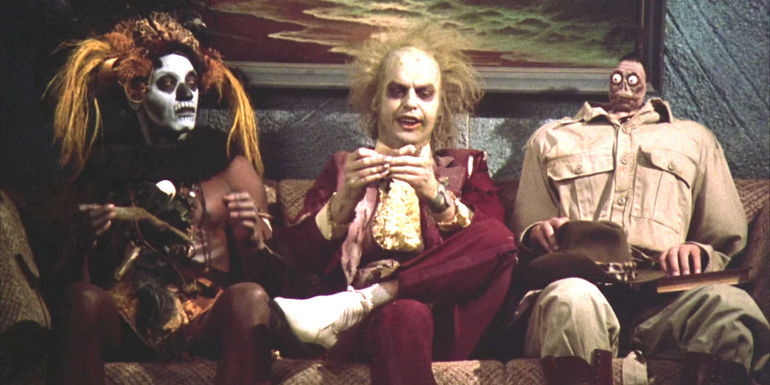 Michael Keaton as Beetlejuice sitting on a sofa between a witch doctor with a white painted face and an explorer with a shrunken head