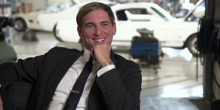 Leo Beebe (Josh Lucas) sitting in chair with his chin on his hand smiling in Ford v Ferrari.