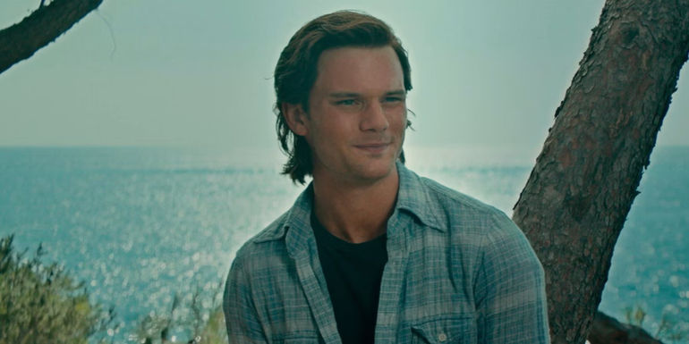 Younger Sam (Jeremy Irvine) leaning against a tree trunk in front of the ocean in Mamma Mia! Here We Go Again.