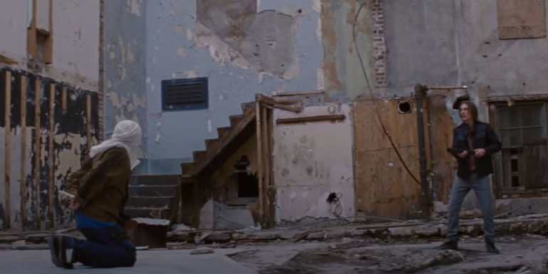 Paul Dano as Seth in Looper aiming a weapon as a kneeling, hooded figure