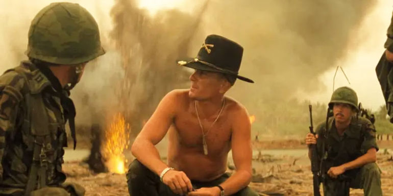 A shirtless Bill Kilgore (Robert Duvall) kneeling down between two soldiers on a battlefield in Apocalypse Now