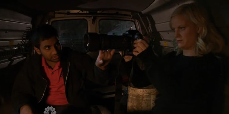Tom looks at Leslie as she tries to use a camera while sitting in a van in Parks and Recreation.