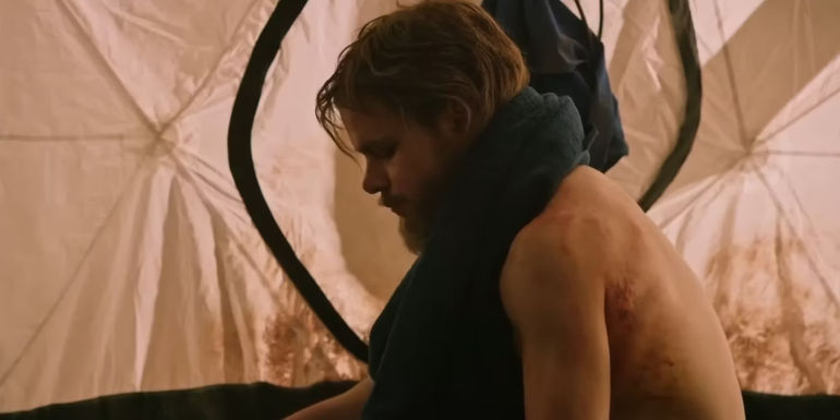 Mark Watney (Matt Damon) with his shirt off showing scars, and eating alone in a tent in The Martian.