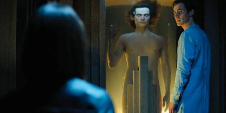 Cassian Bilton as Brother Dawn in Foundation near a tank with a clone of himself inside