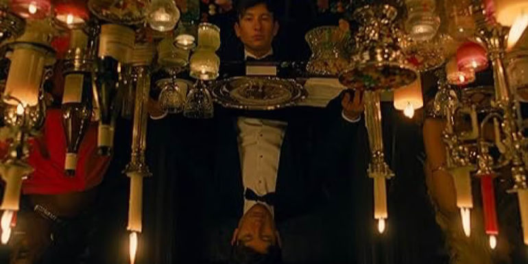 Oliver sits at a lavish dining table which is reflecting his face in Saltburn.