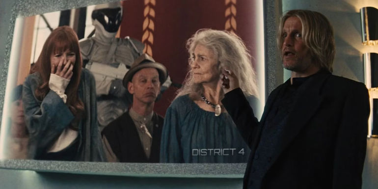 Haymitch points to a screen that shows Mags volunteering for Annie in District 4 in The Hunger Games Catching Fire
