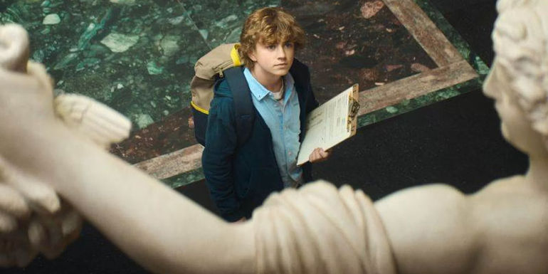 Walker Scobell as Percy Jackson looks up at a statue with a clipboard in his hand in Percy Jackson and the Olympians.