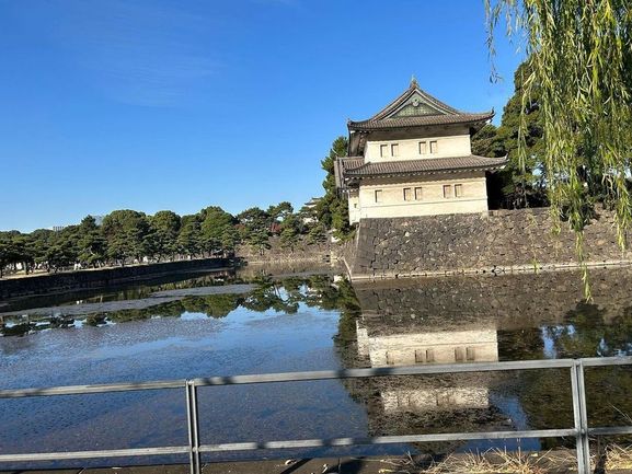 A traditional Japanese building with a tiled roof is reflected in a calm pond, with trees and a clear sky in the background. No people visible.