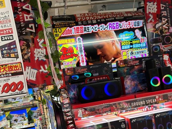 The image shows a cluttered electronics store with colorful advertising signs and speakers on display. There are no people visible.