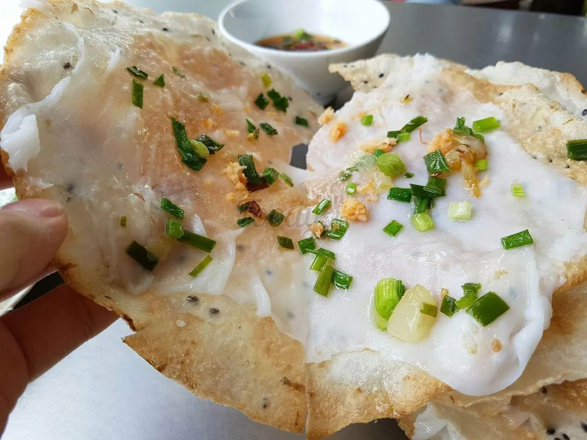 Bánh đập cầm trên tay. Bánh tráng nướng mè phủ lớp bánh ướt, rắc hành lá và tóp mỡ
