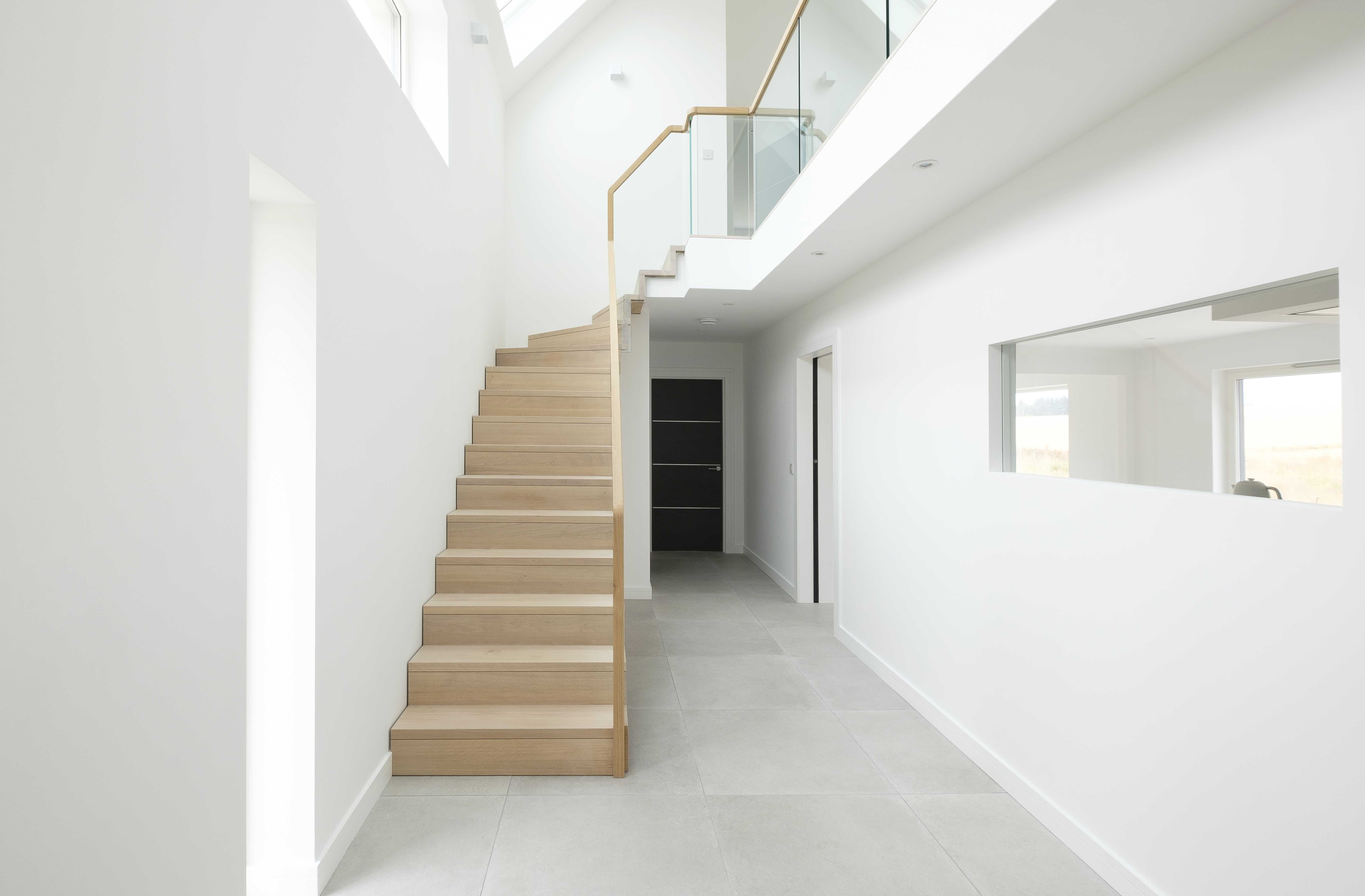 Modern Raw oiled oak stair with glass balustrade in large, minimal, white hall with grey tiles floor and black doors.