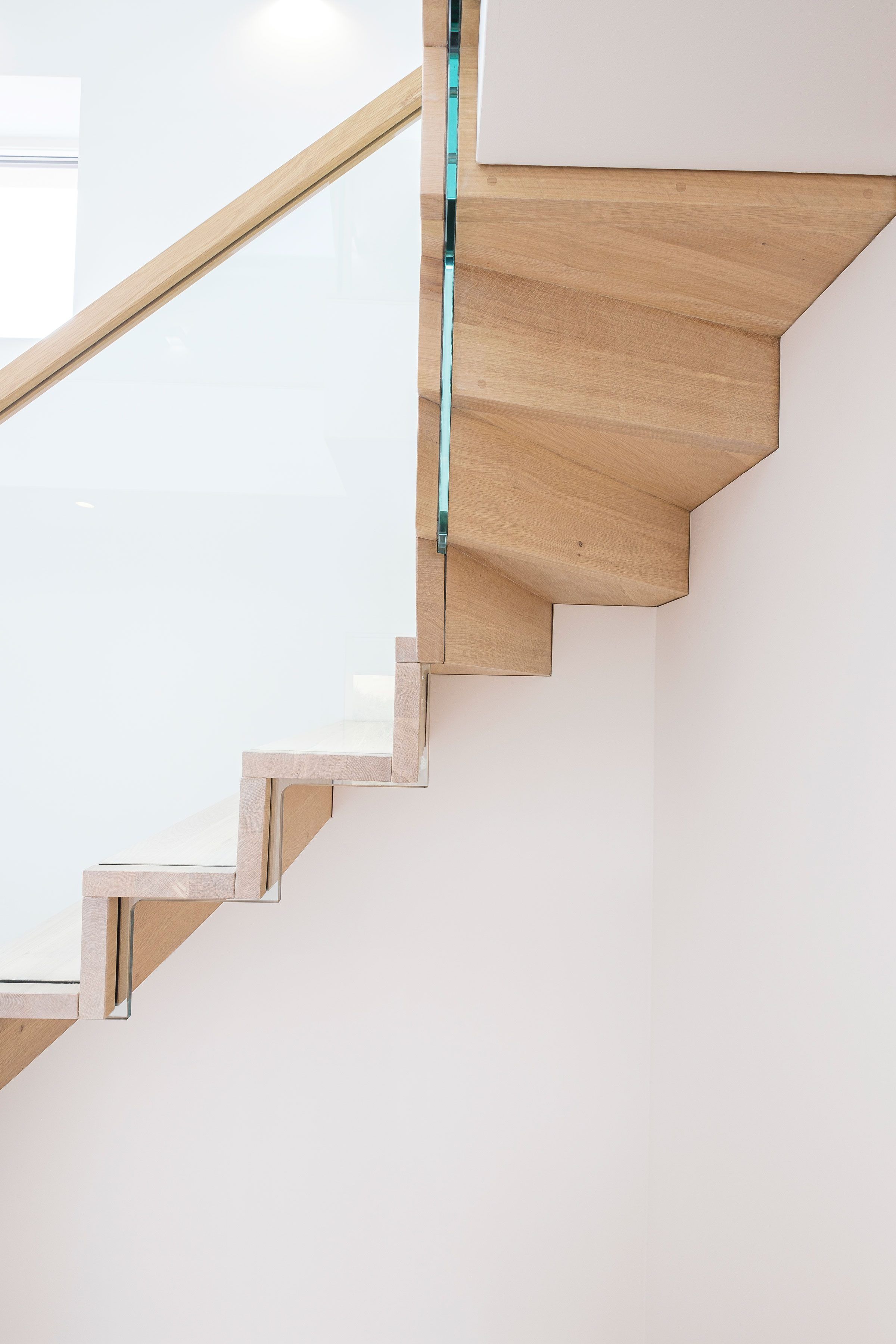 Underside view of slim profile cantilevered oak staircase handrail and glass balustrade. White wall.