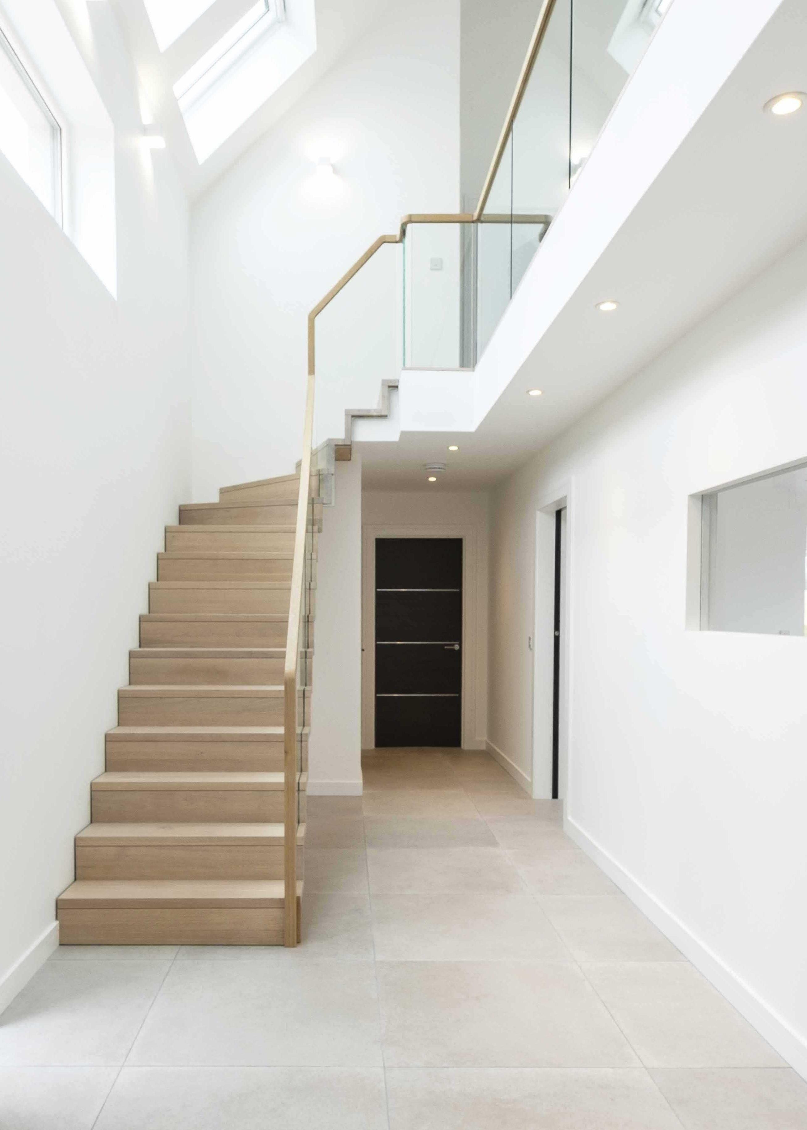 Modern pale oak staircase with glass balustrade in large, minimal, white hall with grey tiles floor and black doors.