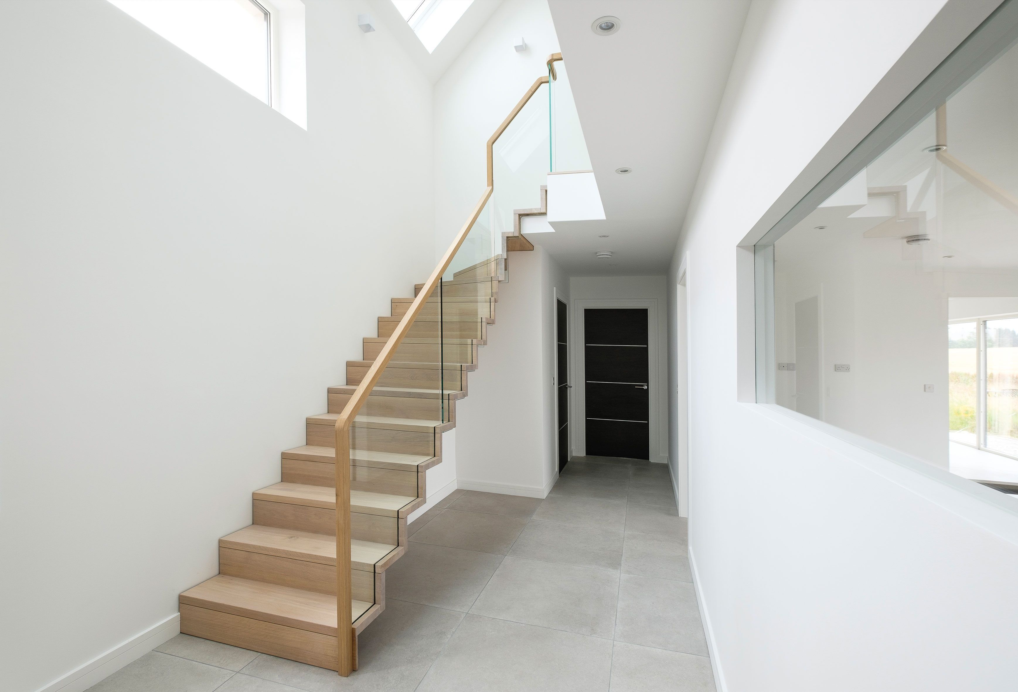 Contemporary bespoke oak staircase with glass balustrade in large, minimal, white hall with grey tiles floor and black doors.