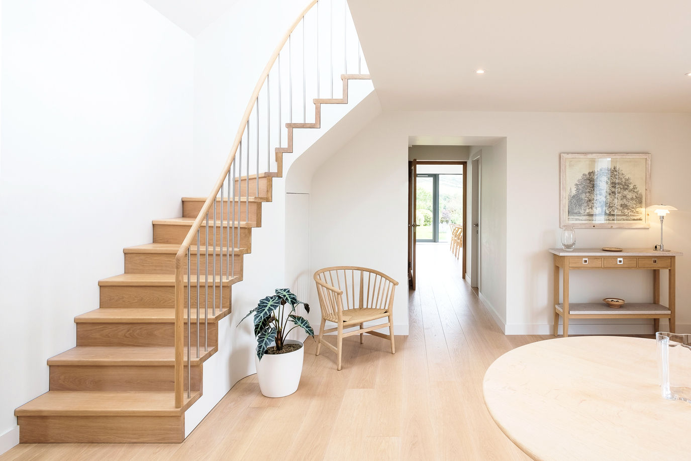 Oak domestic interior with Danish designed oak table and chair, oak staircase and handrail with stainless steel balustrade. White walls and potted plant.