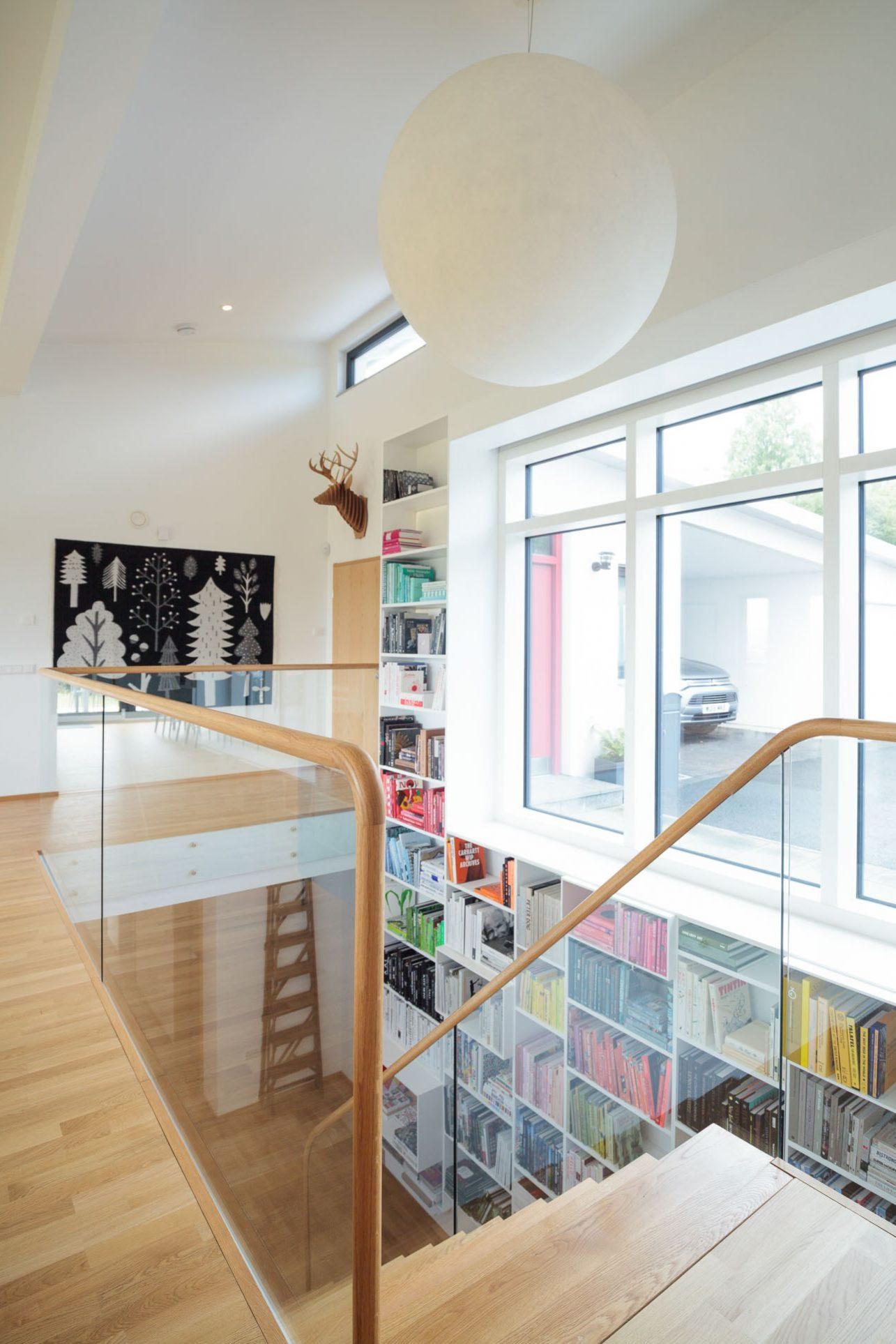 Glass balustrade of staircase with oak handrail. White built-in bookcases with rows of books organised by colour. Black illustrated tapestry on wall and paper lantern light shade.