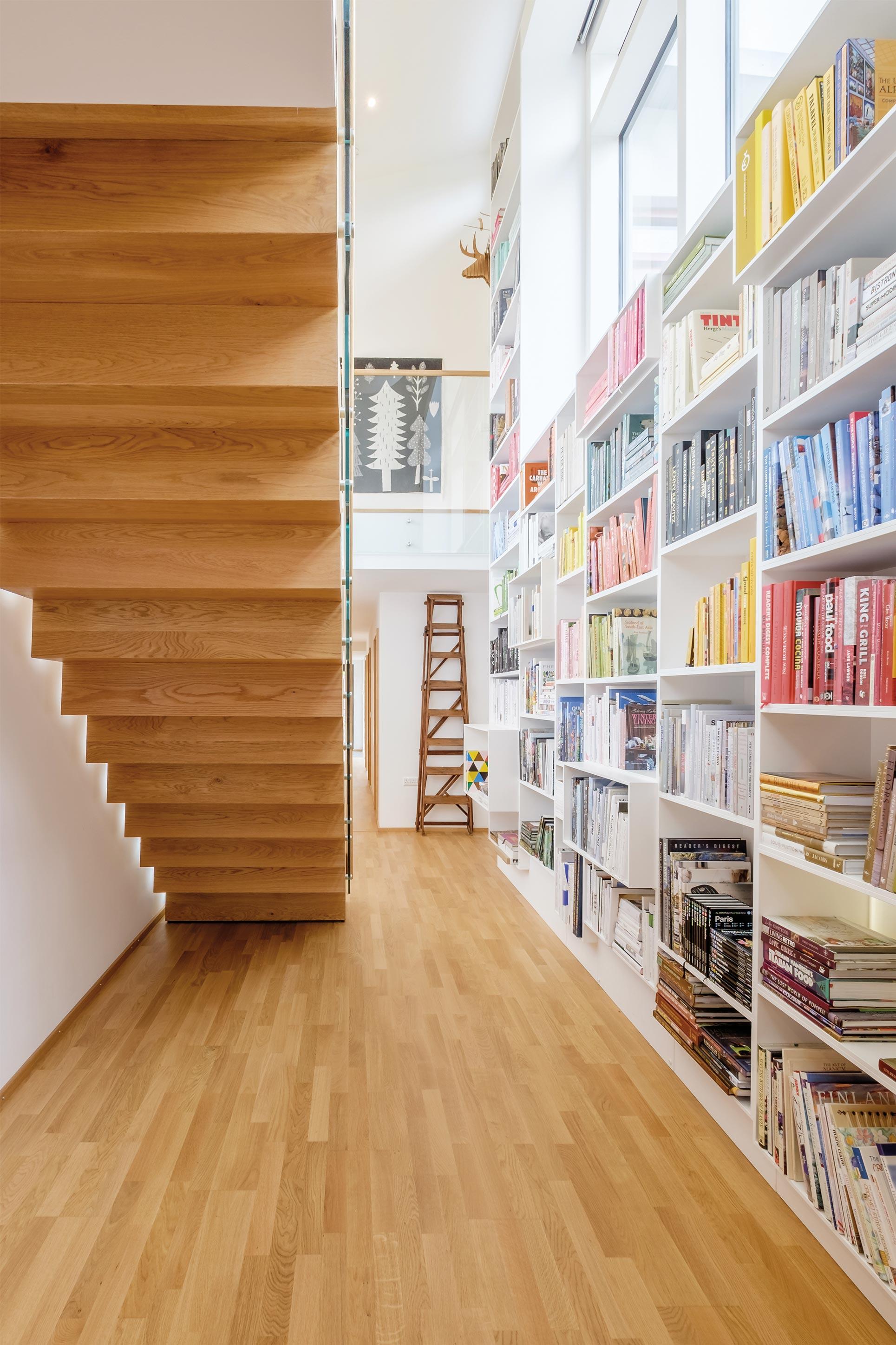 Underside of sculptural oak zig zag staircase. White fitted bookcases with rows of books organised by colour.