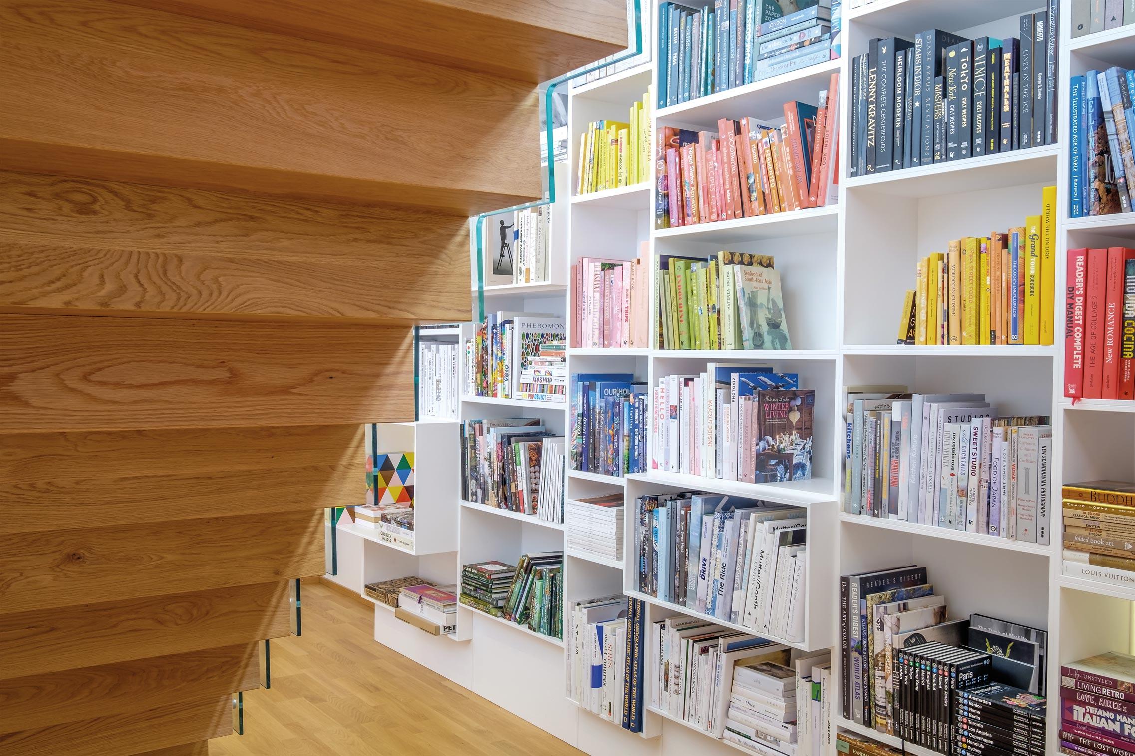 Underside of sculptural oak zig zag stair. White fitted bookcases with rows of books organised by colour.