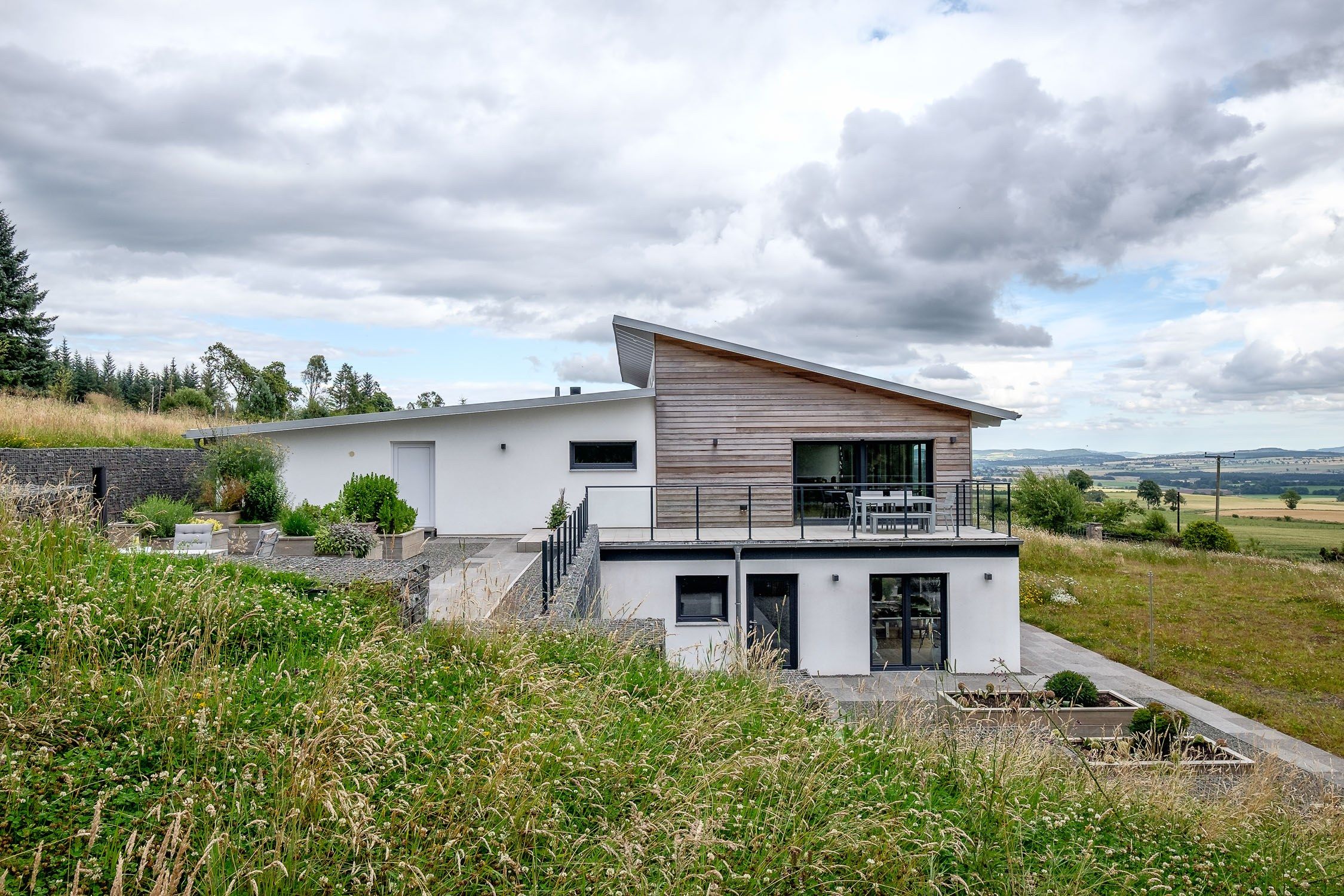 Contemporary designed home with off set roofs, larch cladding, white render, Gabion baskets and green garden.