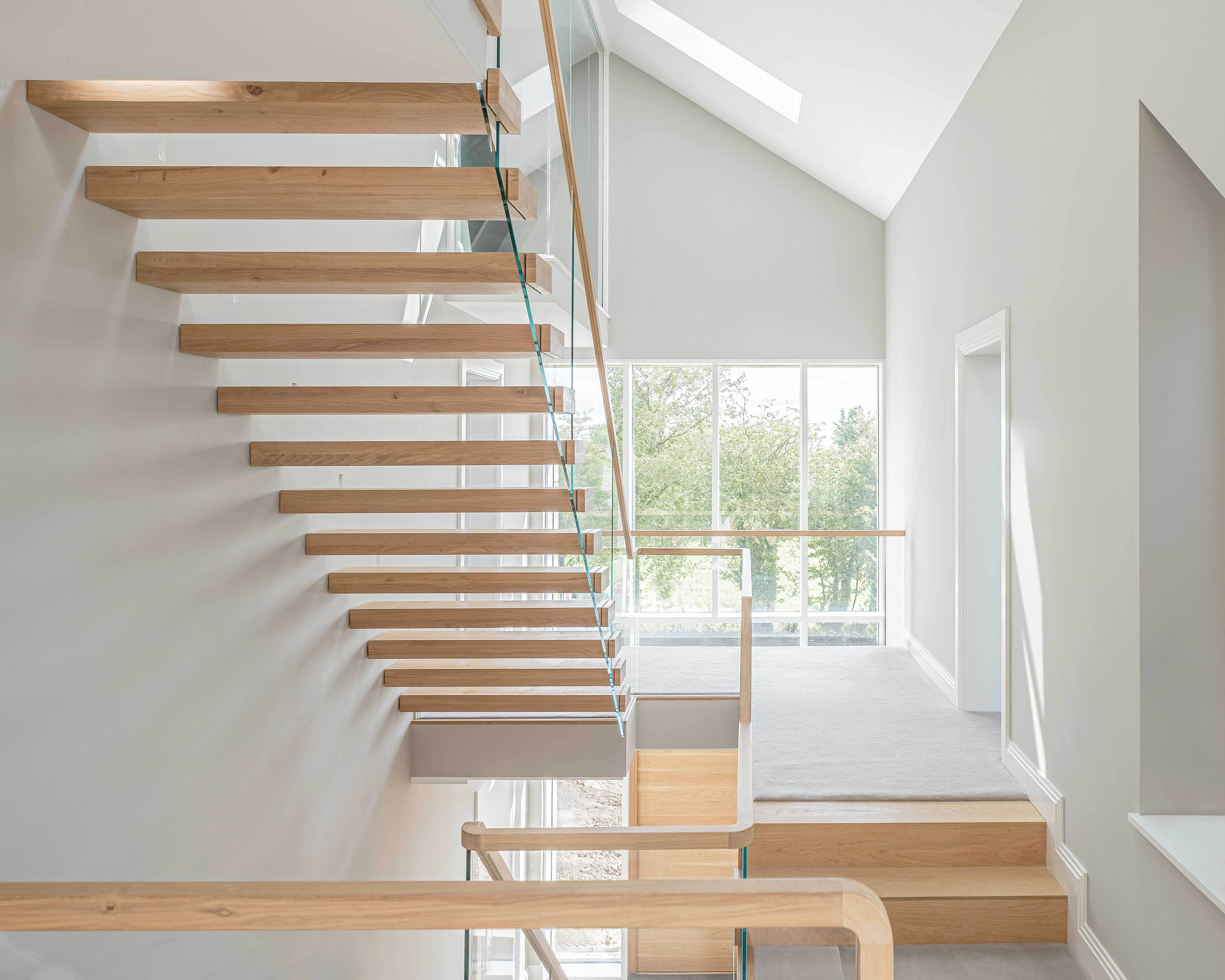 Underside view of cantilevered floating oak stair with glass balustrade from first floor. Grey walls and carpet.