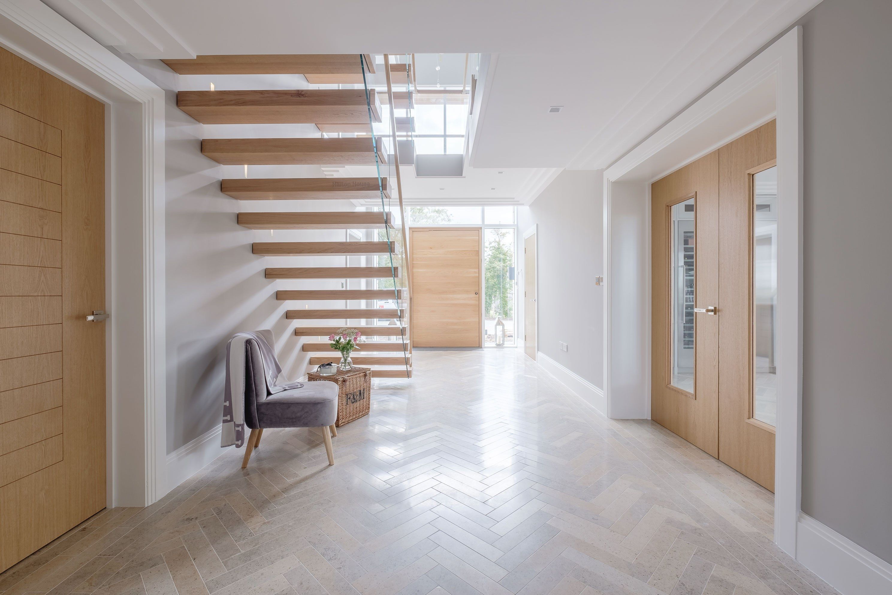 Underside of cantilevered oak stair with glass balustrade from ground floor level. Grey upholstered chair and basket under stair. Wooden doors and herringbone tiled flooring.