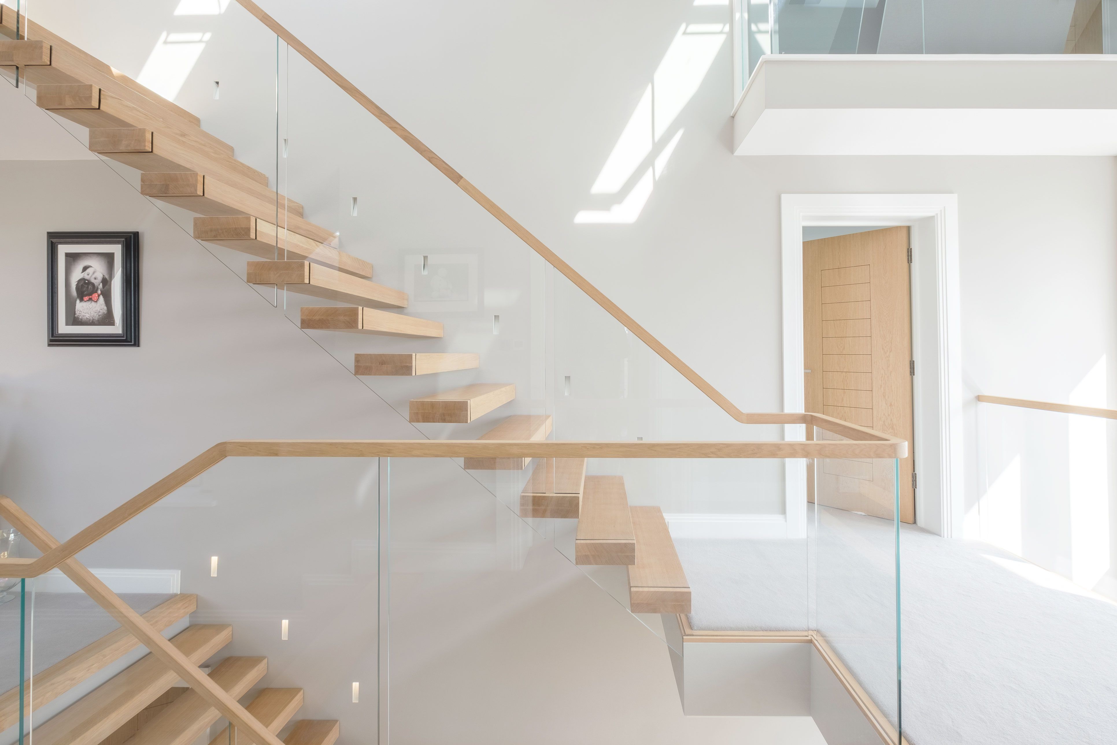 Side view of cantilevered floating oak stair over three stories with glass balustrade. Grey walls and carpet with oak doors.